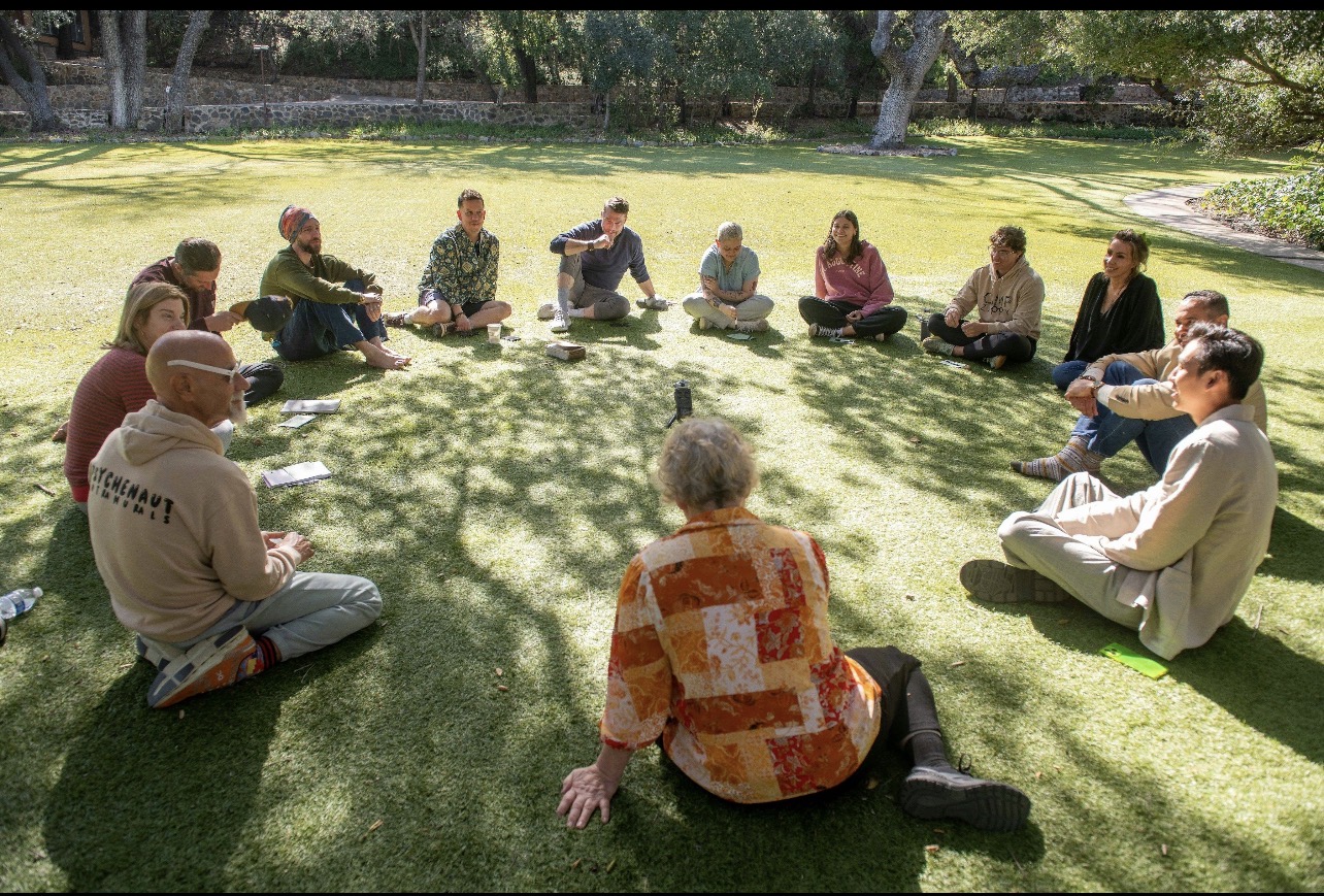 A circle of people seated together on grass beneath oak trees